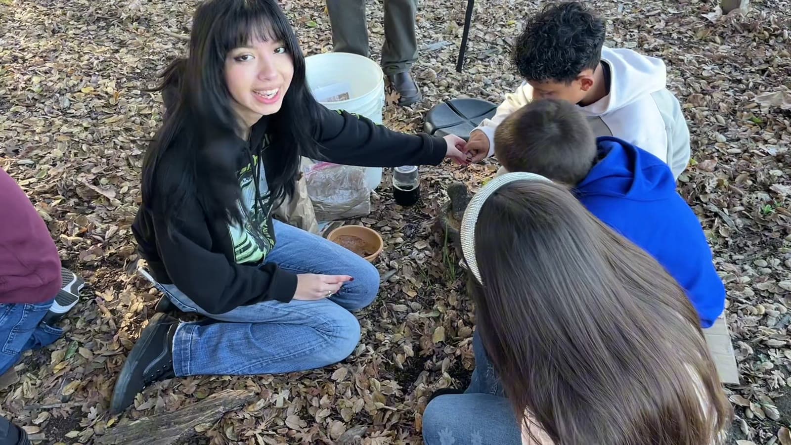 A Druids docent, smiling, works with younger students around a grinding bowl on the forest floor