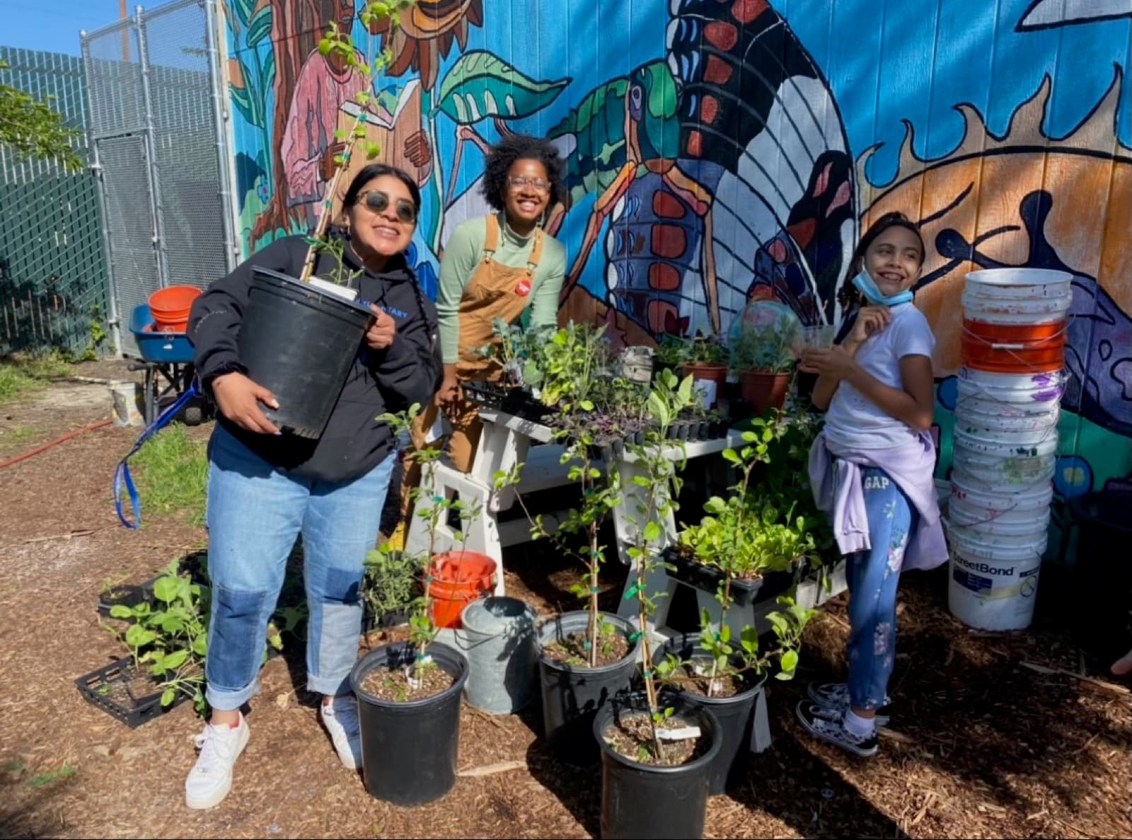 Volunteers smile beside potted tree saplings and trays of plant starts in front of a large painted butterfly mural.