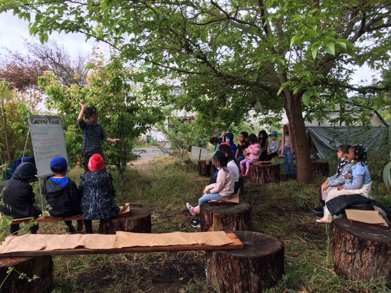 An educator points to a whiteboard titled Garden Rules while a class sits on log rounds under a tree.