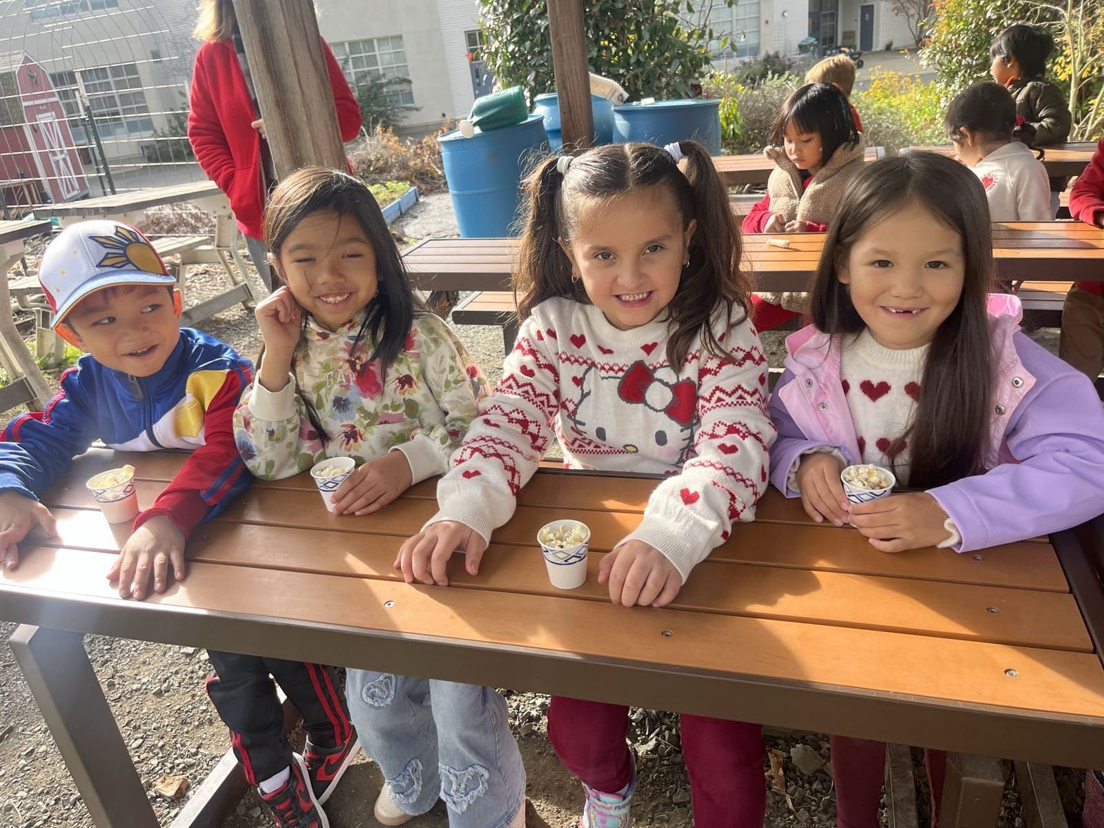 Four young students smile at an outdoor picnic table, each holding a small paper cup of a snack.