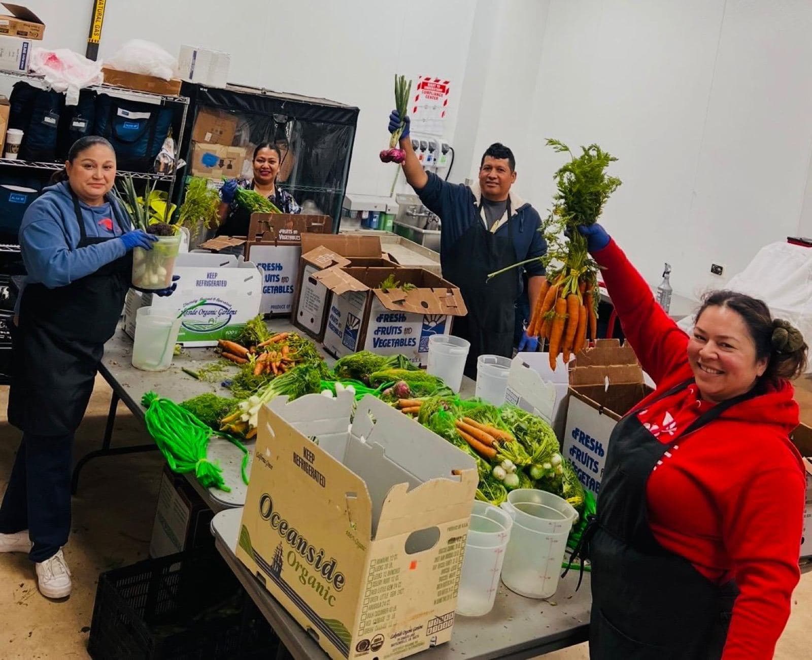 Four workers in aprons and gloves hold up bunches of carrots and green onions over boxes of produce.