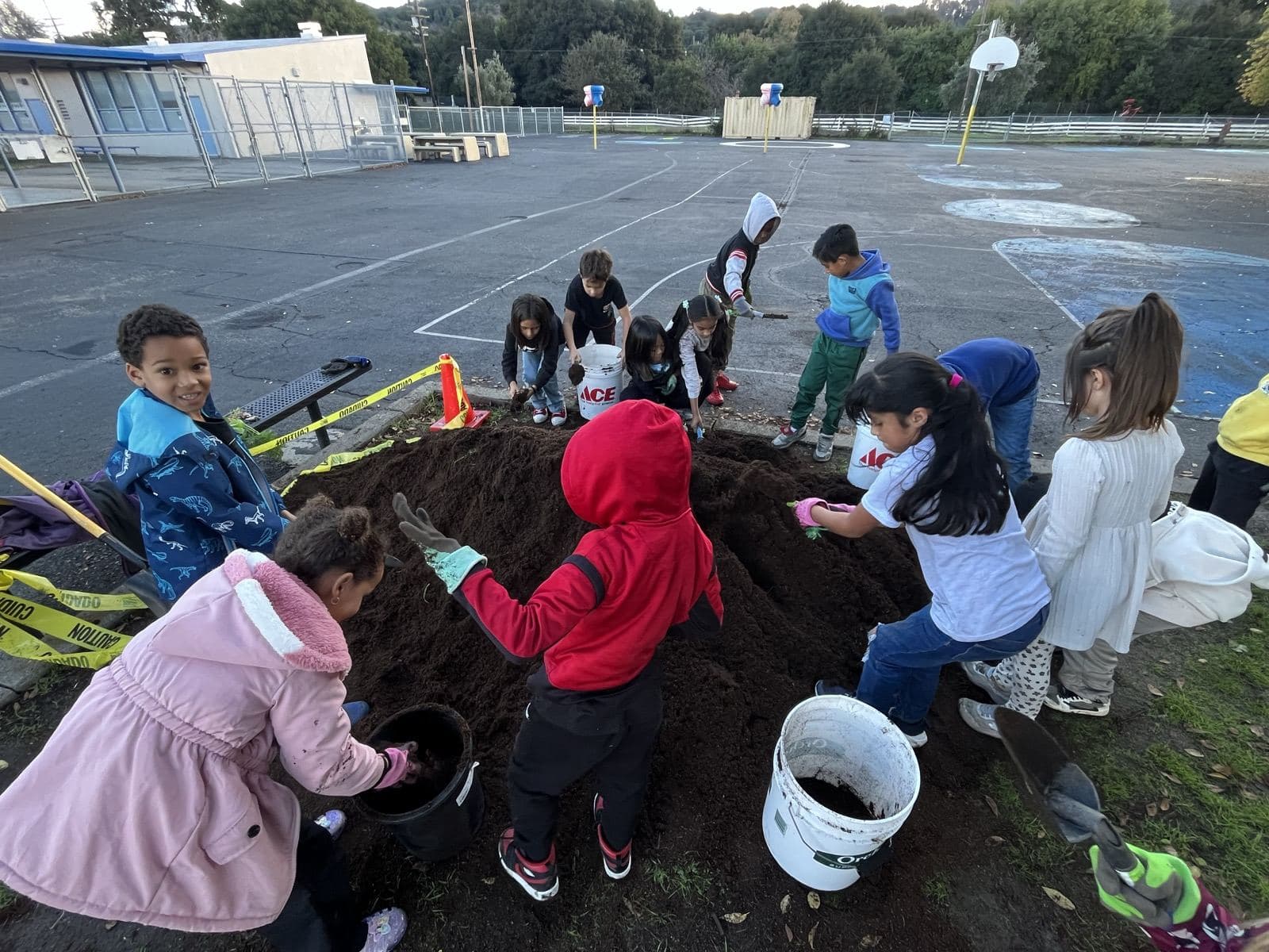 Children scoop dark soil from a large mound into buckets beside a blacktop basketball court, caution tape strung nearby.