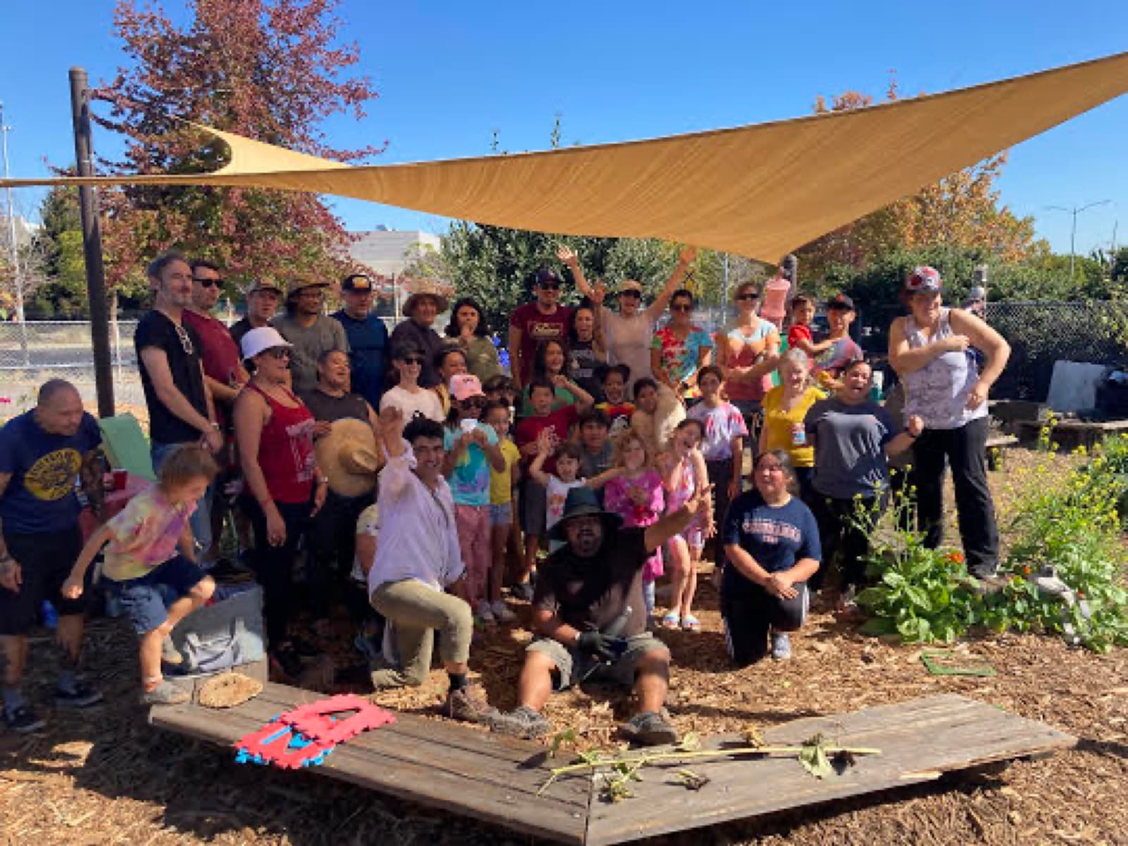 A large group of children and adults cheers together under a tan shade sail in a mulched school garden.