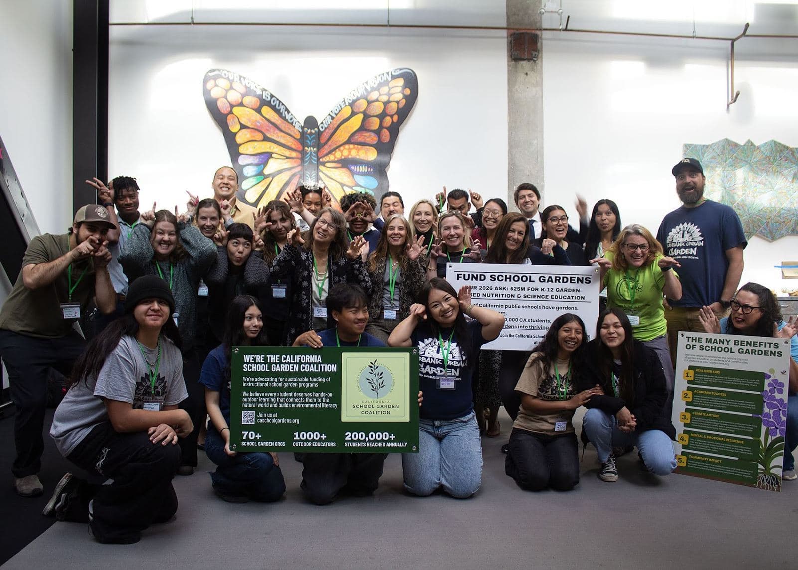 A large group poses under a painted monarch butterfly with banners reading California School Garden Coalition and Fund School Gardens.