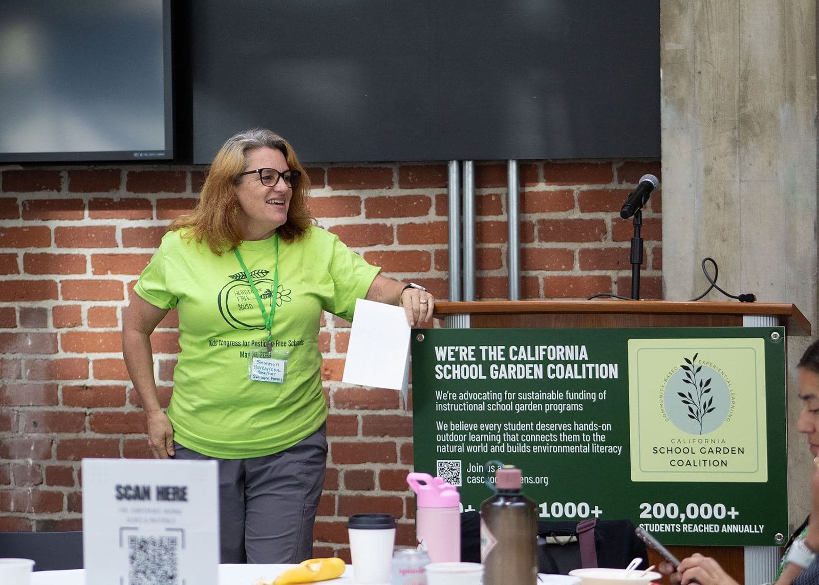 A woman in a green shirt speaks at a podium hung with a California School Garden Coalition banner.