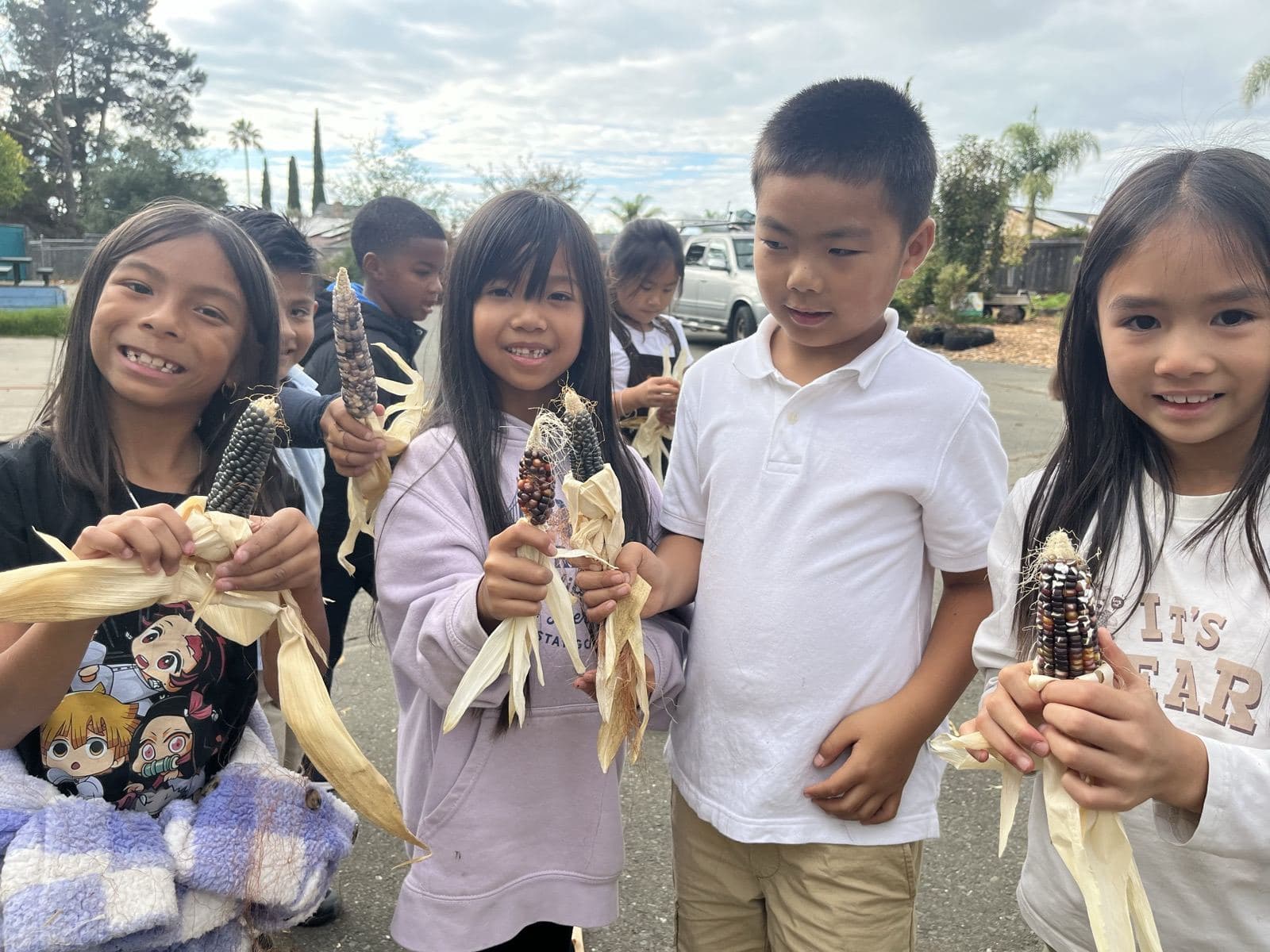 Students grin and hold up ears of speckled flint corn, husks peeled back.