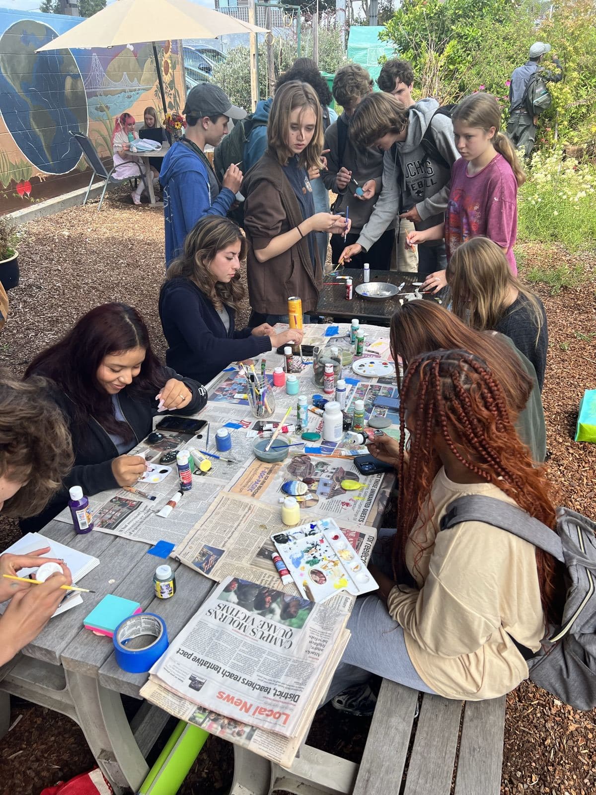 Students crowd around a newspaper-covered picnic table, painting rocks beside a garden mural.